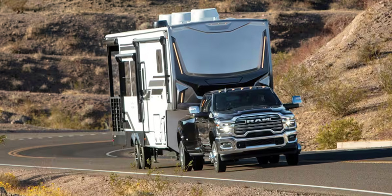A RAM 3500 towing a large RV trailer on a winding road through a desert landscape.