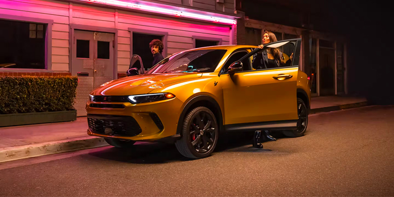Night street scene featuring a gold/yellow SUV with the driver’s door open under neon pink lighting, with a woman entering the vehicle and a man standing nearby outside a building.