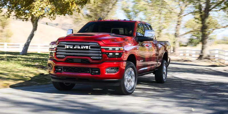Front three-quarter view of a red RAM pickup truck driving on a tree-lined road in daylight, showing motion and chrome grille details.