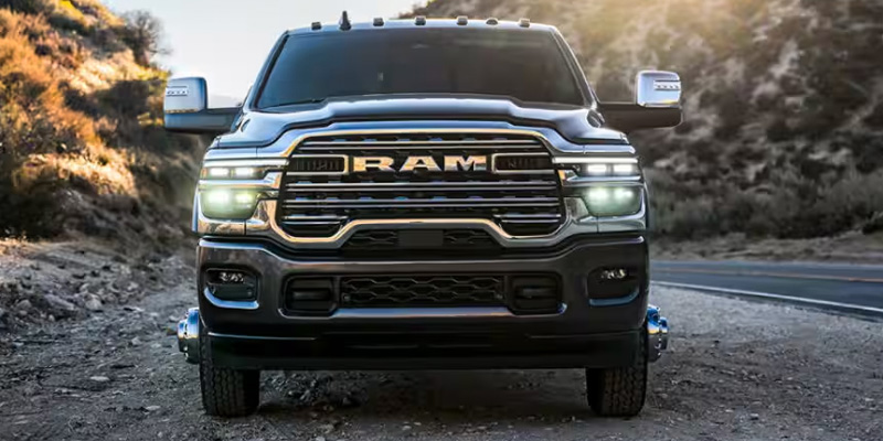 Front view of a black RAM heavy-duty truck driving on a canyon road, with headlights on and rugged terrain in the background.