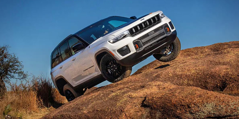 Front three-quarter view of a white SUV climbing a steep rocky off-road incline, with one wheel lifted, showing rugged terrain capability and adventure driving conditions.