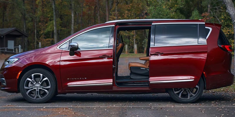 Red Chrysler Pacifica minivan with sliding doors open parked in a wooded outdoor setting.